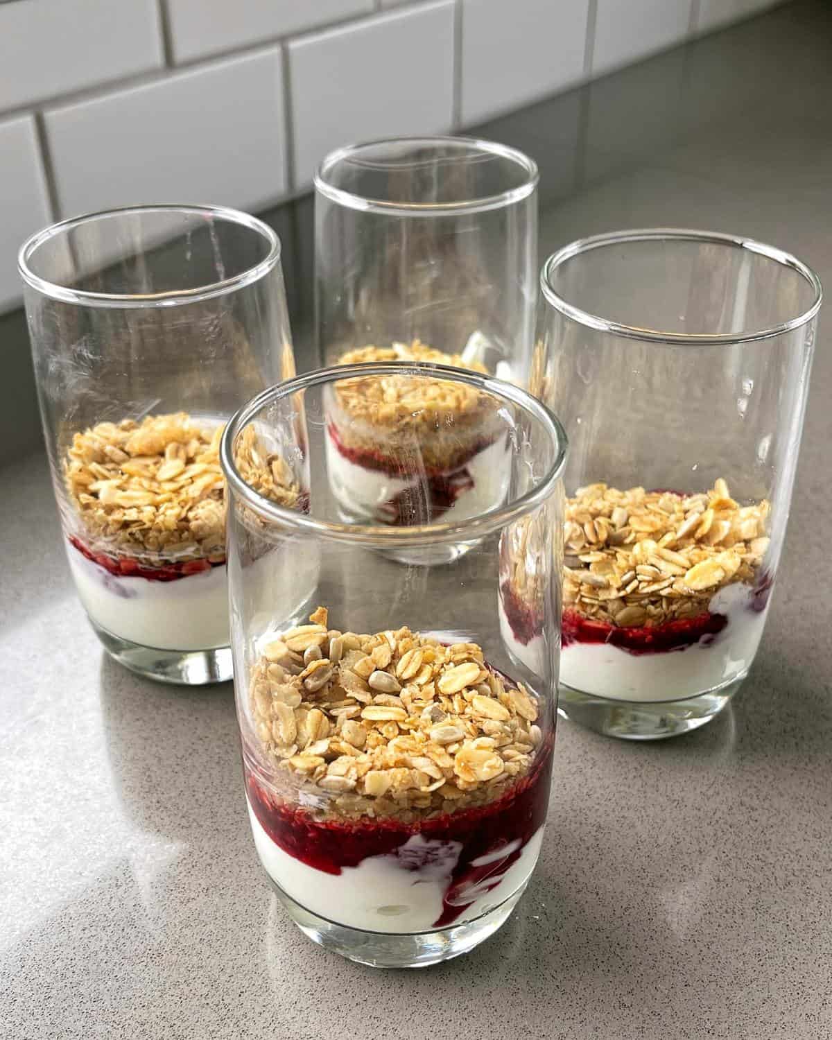 4 tall glasses layered with yoghurt, berry compote and toasted muesli on a grey bench.