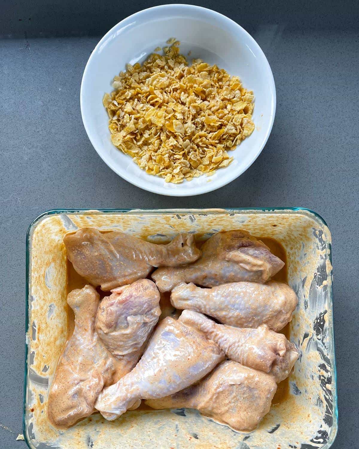 A bowl of cornflakes and a container of marinated chicken on a grey bench.