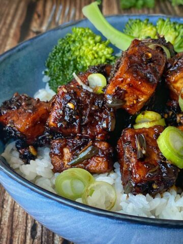 One portion of Black Pepper Tofu served in a blue bowl over steamed rice with broccoli and sliced onions to accompany.