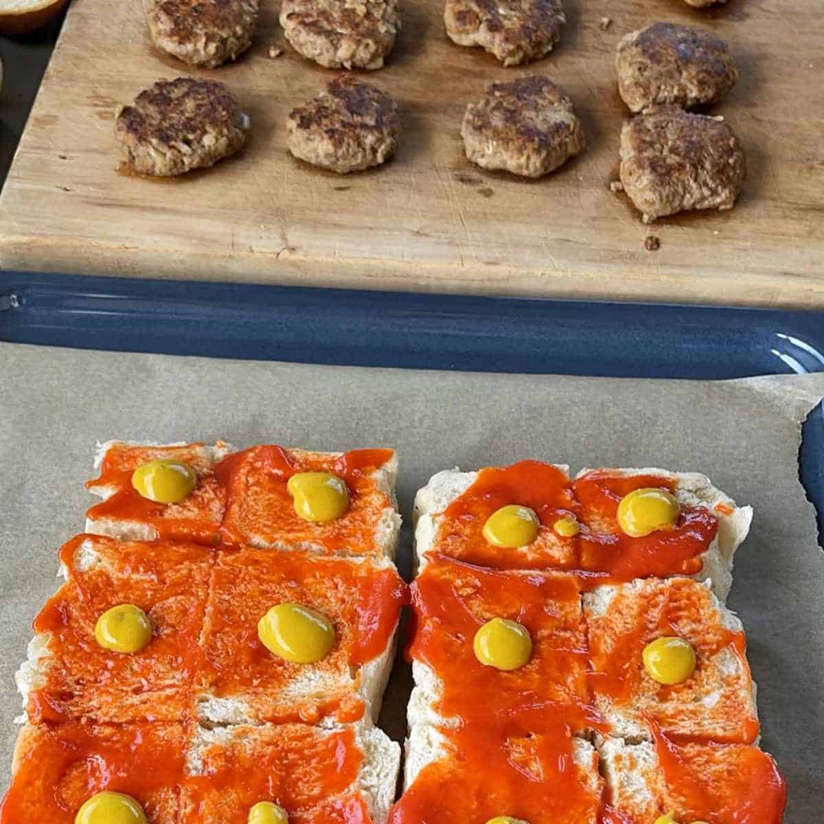 A shot of sliced buns with sauces added and burger patties resting on a wooden chopping board before the Cheeseburger Sliders are assembled and heated through in the oven.