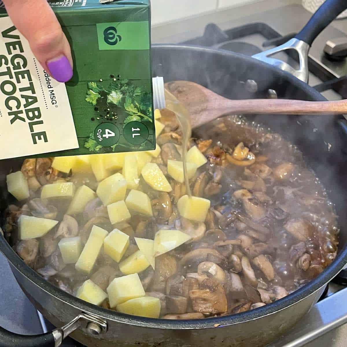 The vegetable stock being added to the sliced mushrooms, garlic, thyme, potatoes and onion in a frypan.