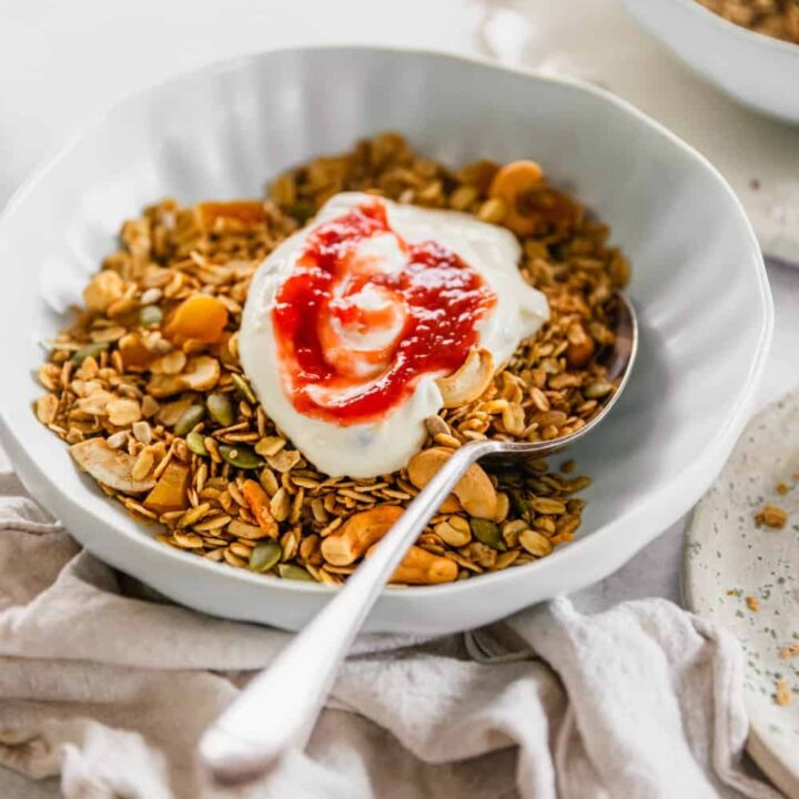 A close up of a bowl of muesli with strawberry yoghurt on top and a spoon in it, sitting on a linen napkin on a white bench.