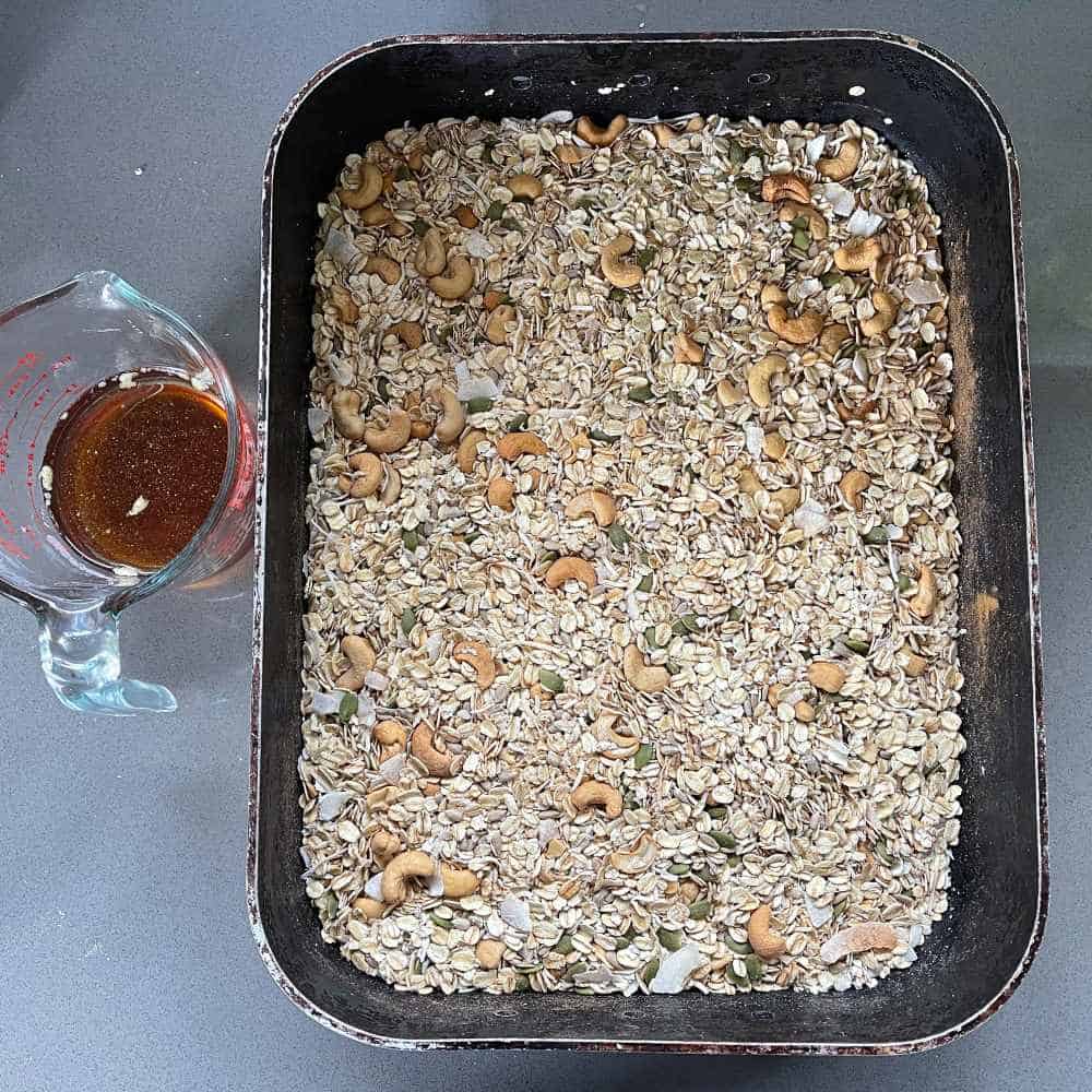 Uncooked muesli in a large roasting dish and a glass jug of oil and maple syrup on a grey bench