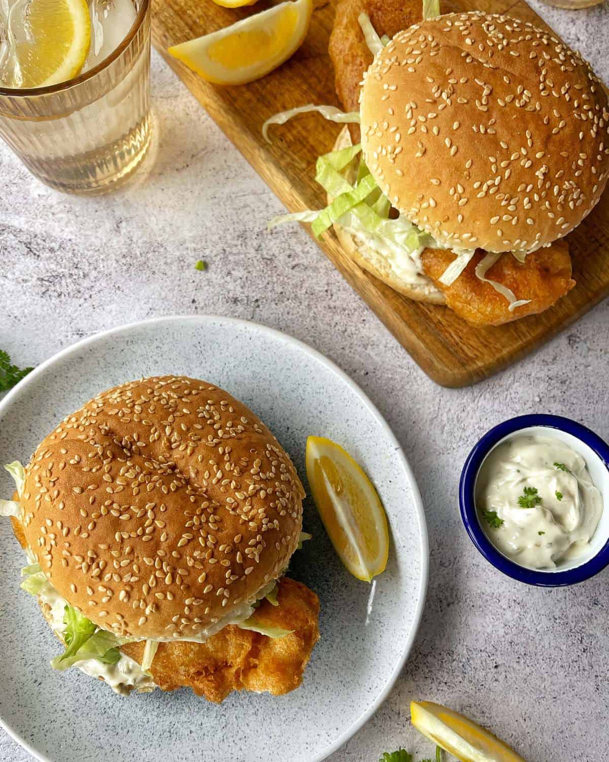 One battered fish burger served on a duckegg blue plate with another burger on a wooden chopping board in the background.