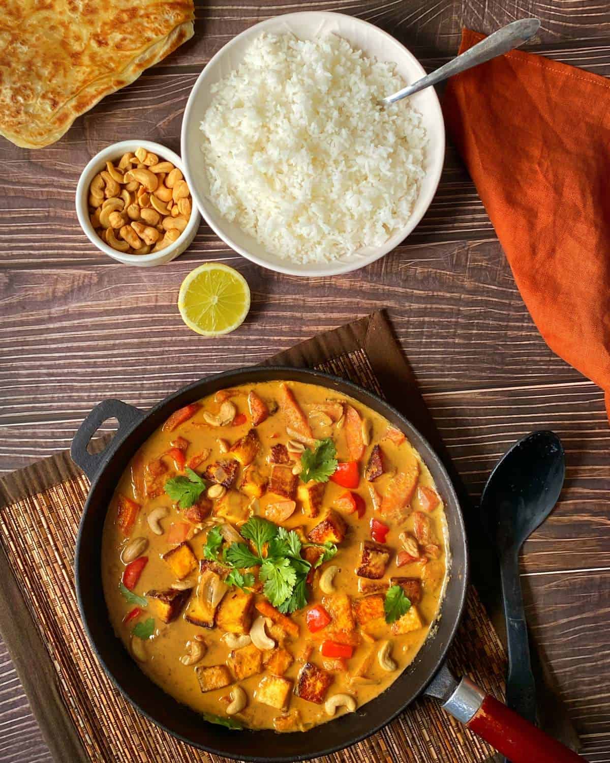 The cooked Paneer Curry garnished with coriander served in the frypan on a wooden table with fluffy white rice, cashew nuts and nan bread to the side.
