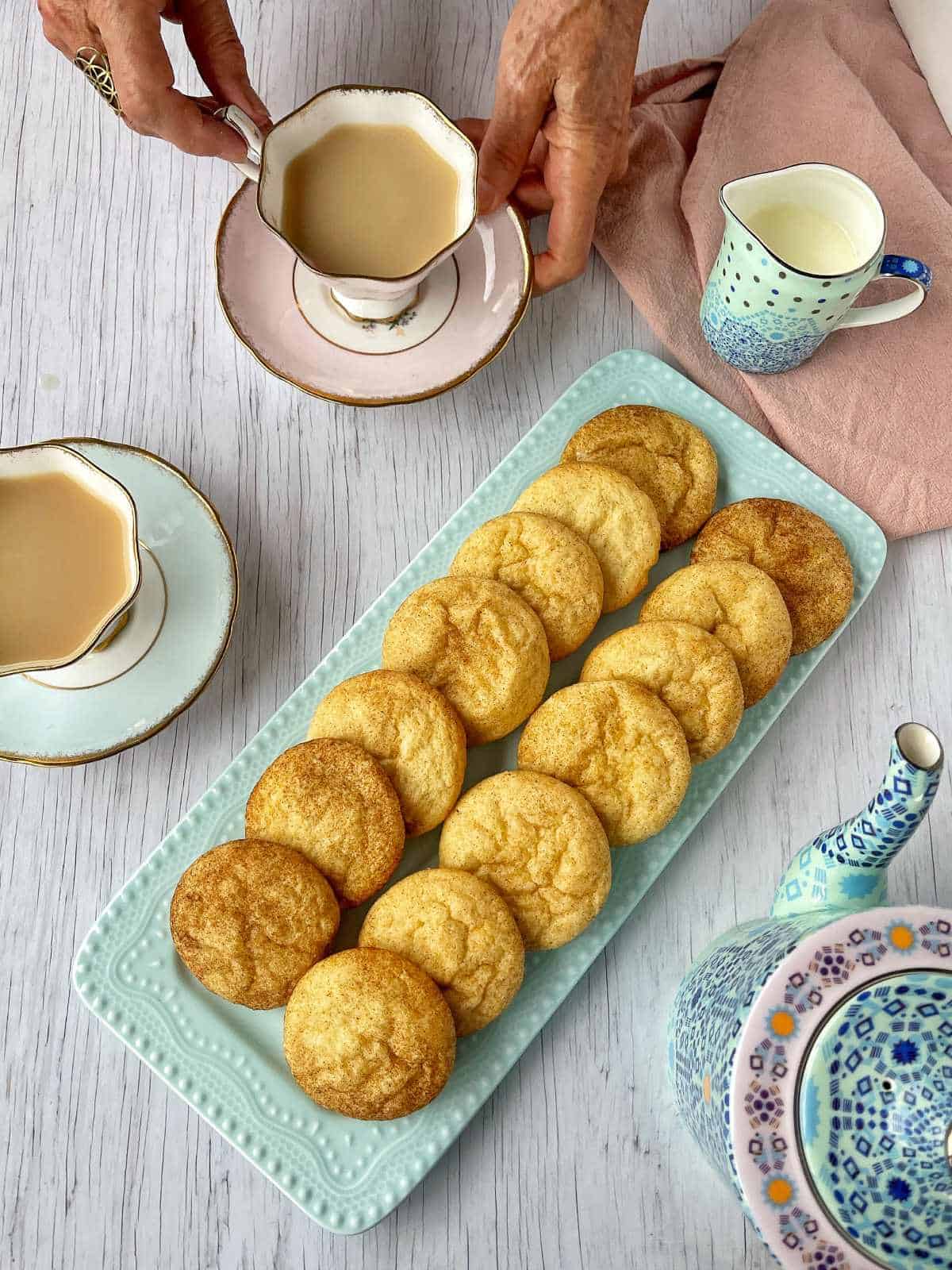 A plate of snickerdoodle cookies in a blue plate on a white bench with cups of tea surrounding it.