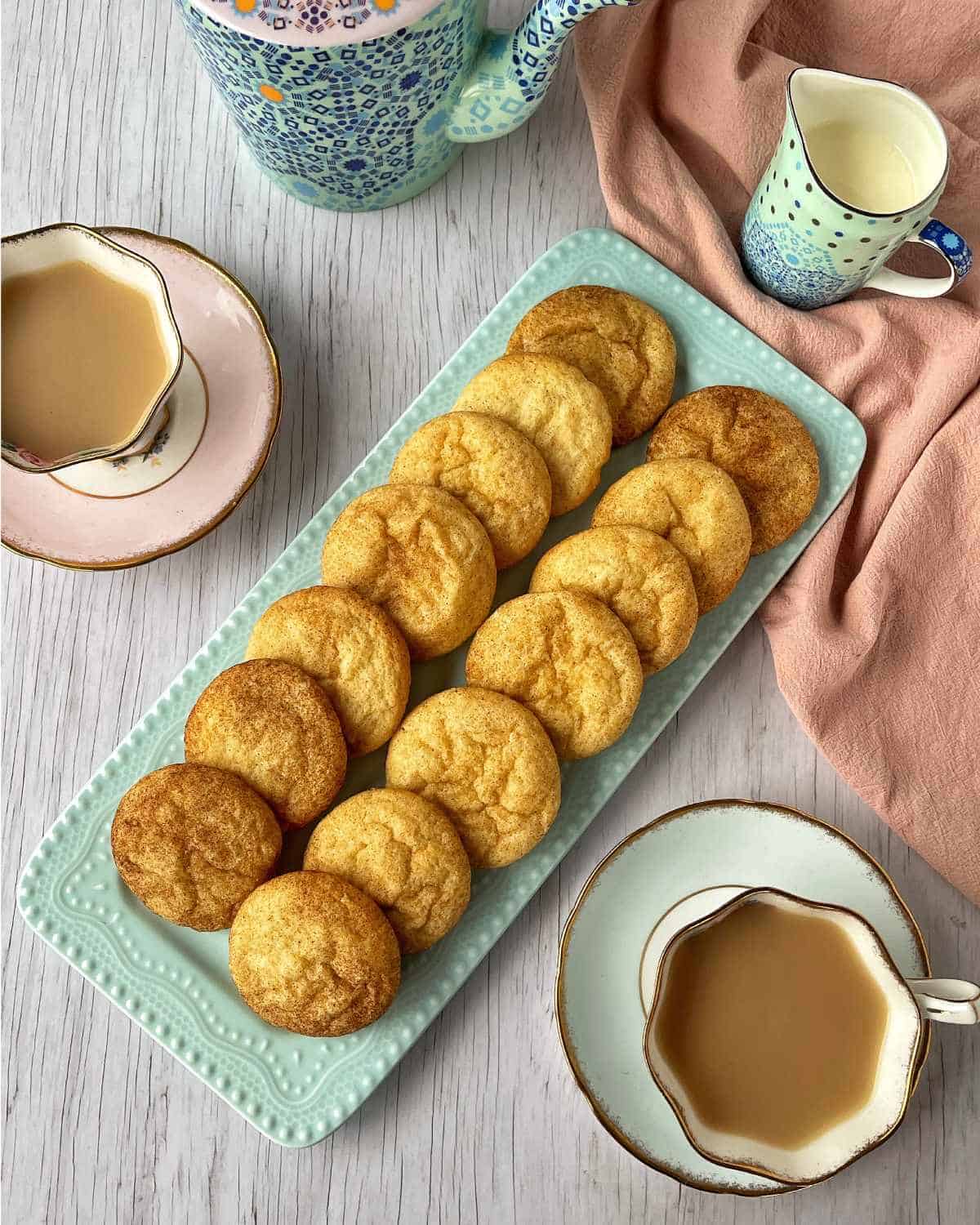 Baked snickerdoodle cookies on a blue platter surrounded by cups of tea.