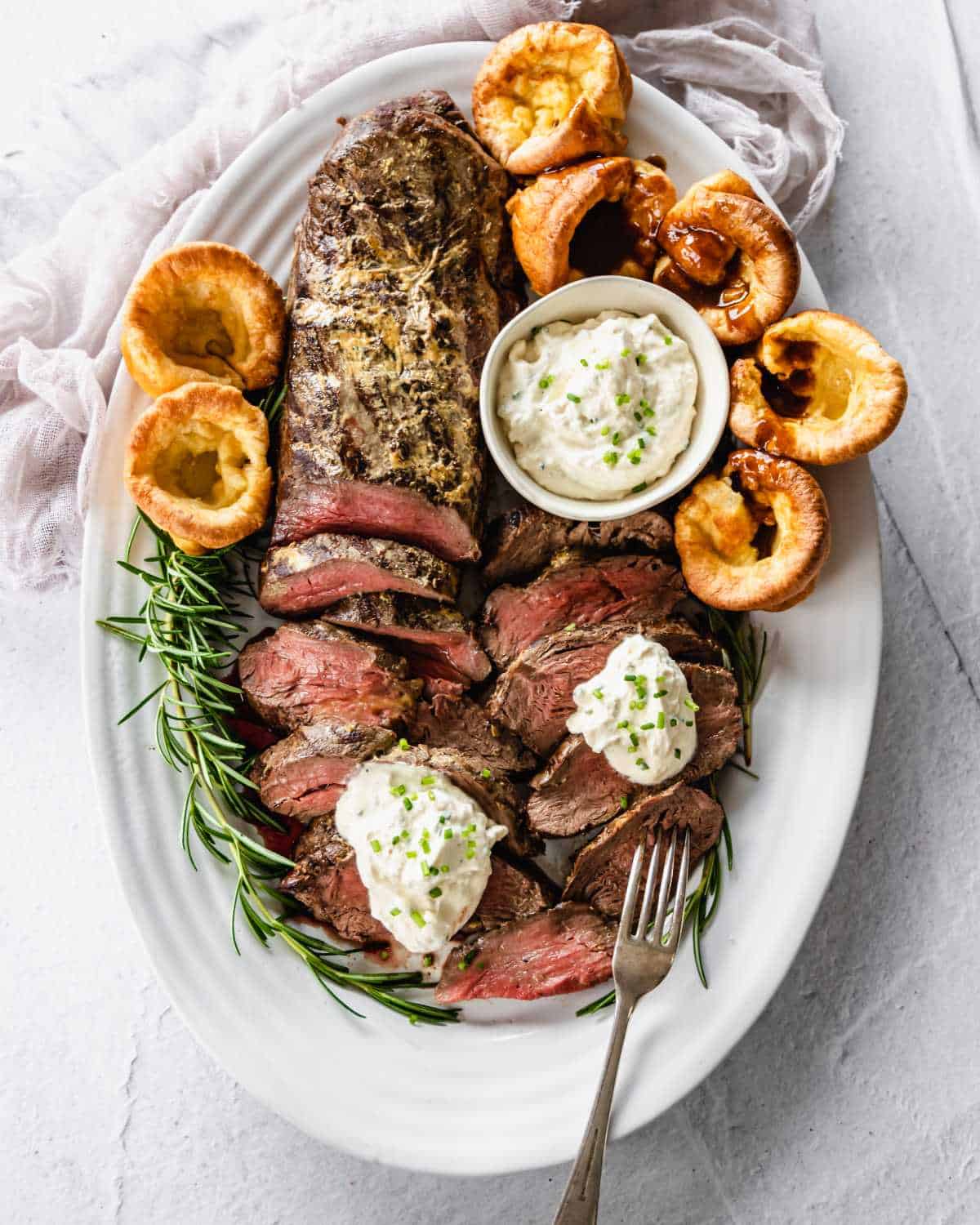 Sliced beef filet on a white serving platter with rosemary and Yorkshire puddings. A small bowl of creamed horseradish to serve.