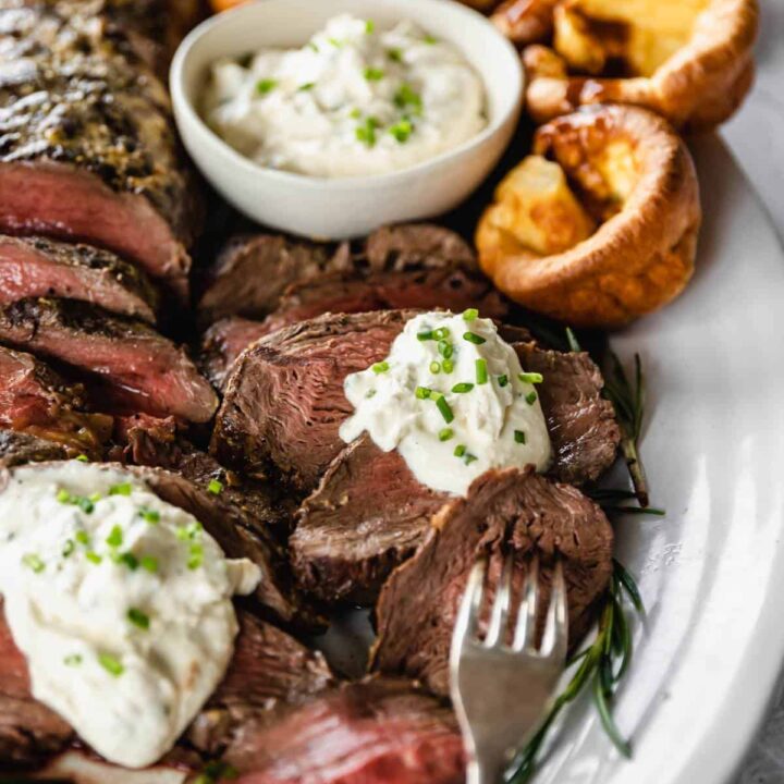 Sliced beef filet on a white serving platter with rosemary and Yorkshire puddings. A small bowl of creamed horseradish to serve.