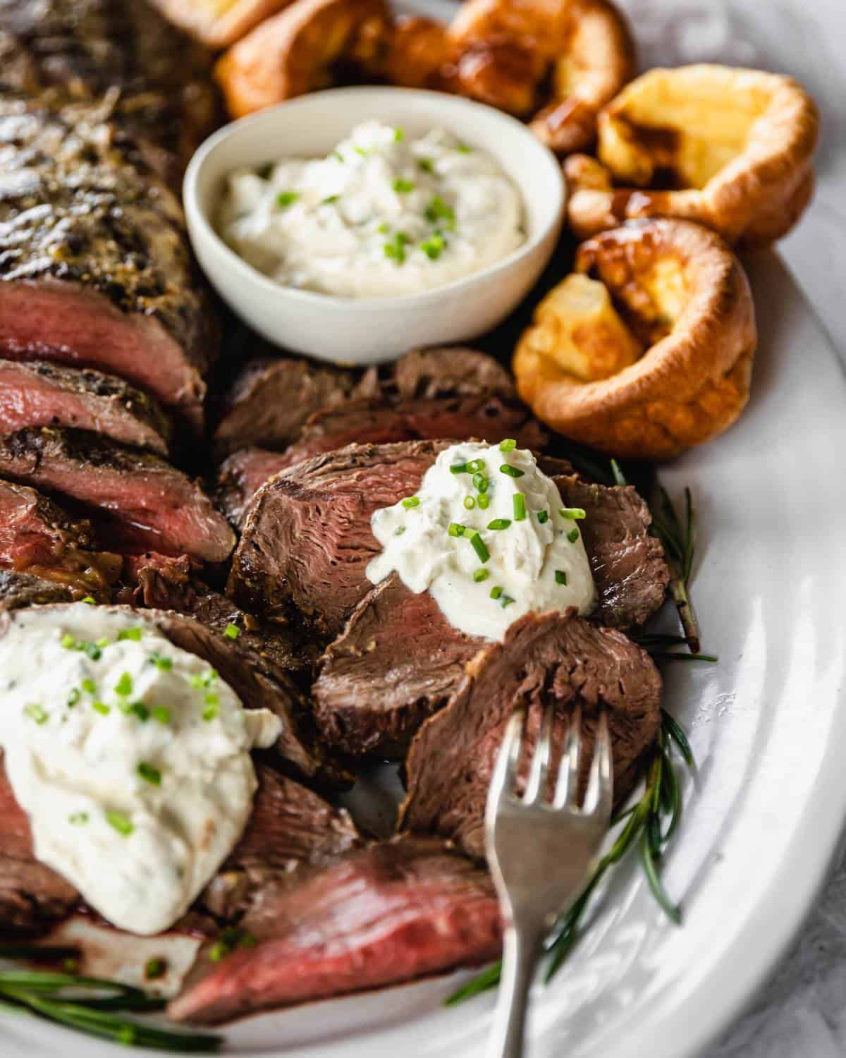 Sliced beef filet on a white serving platter with rosemary and Yorkshire puddings. A small bowl of creamed horseradish to serve.