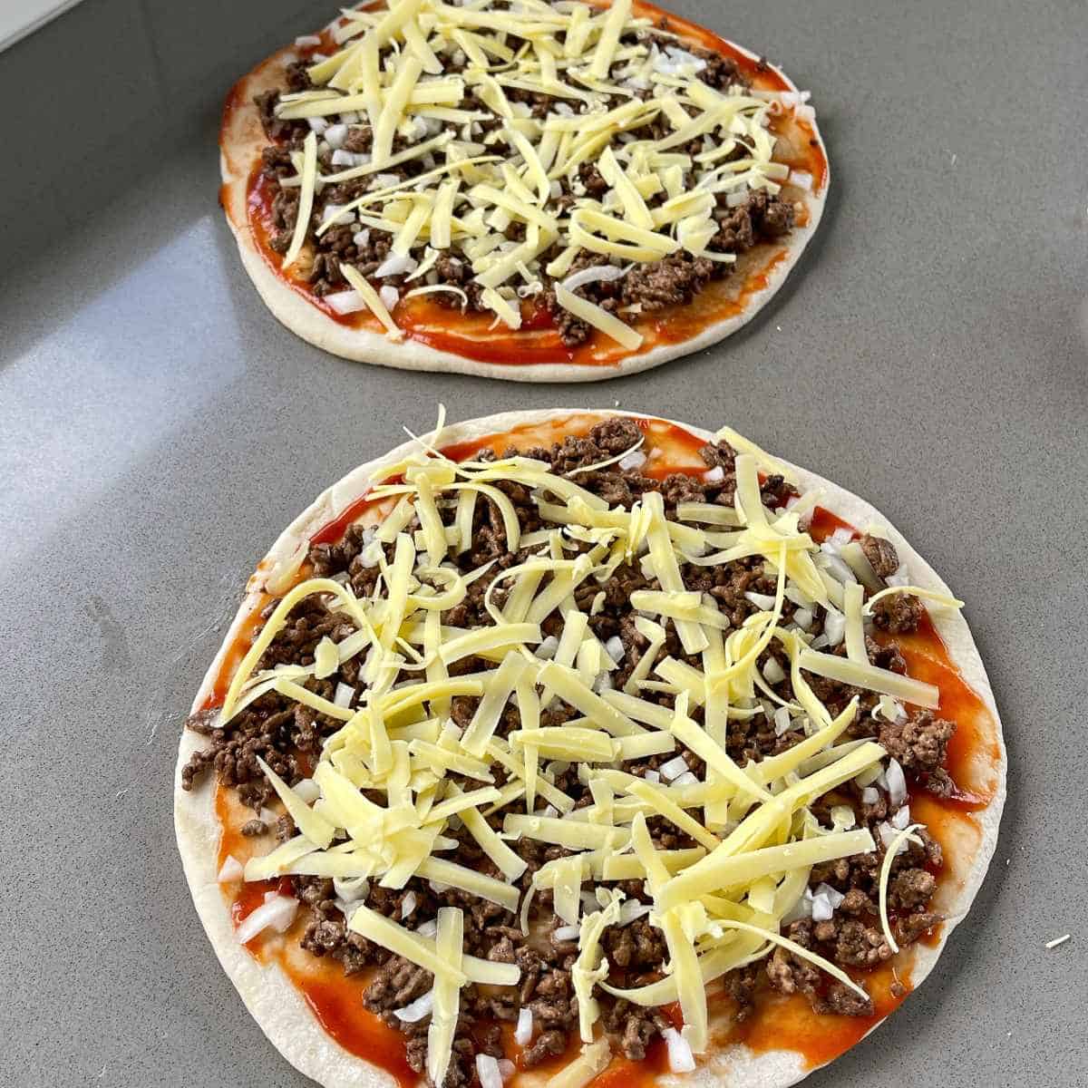Two Big Mac Pizzas with the base sauce, mince, diced onion and grated cheese on them sitting on a grey bench tiop.