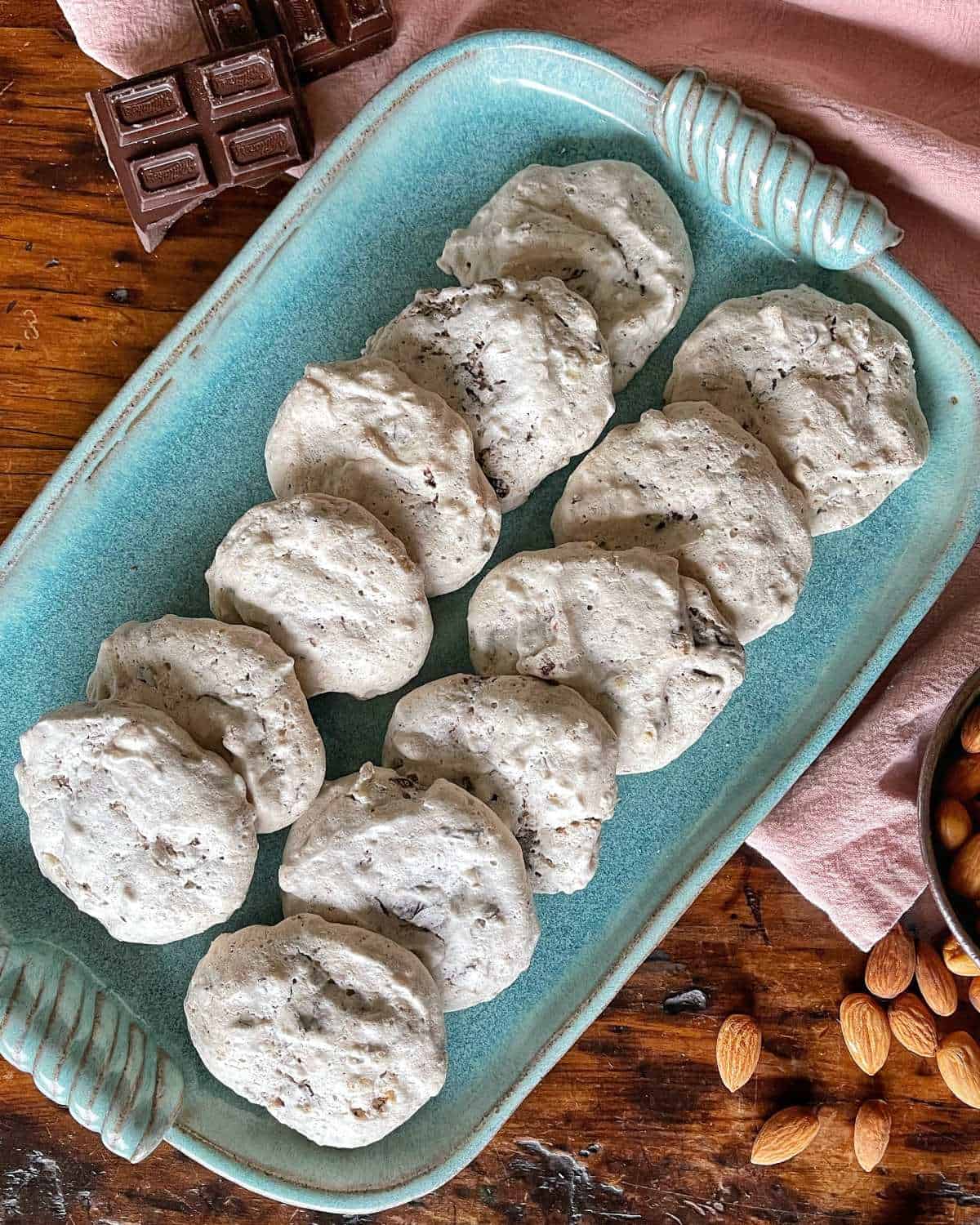 Chocolate Almond Meringue Cookies served on a long blue serving platter on a wooden table with a broken piece of chocolate and scattered almonds to the side.