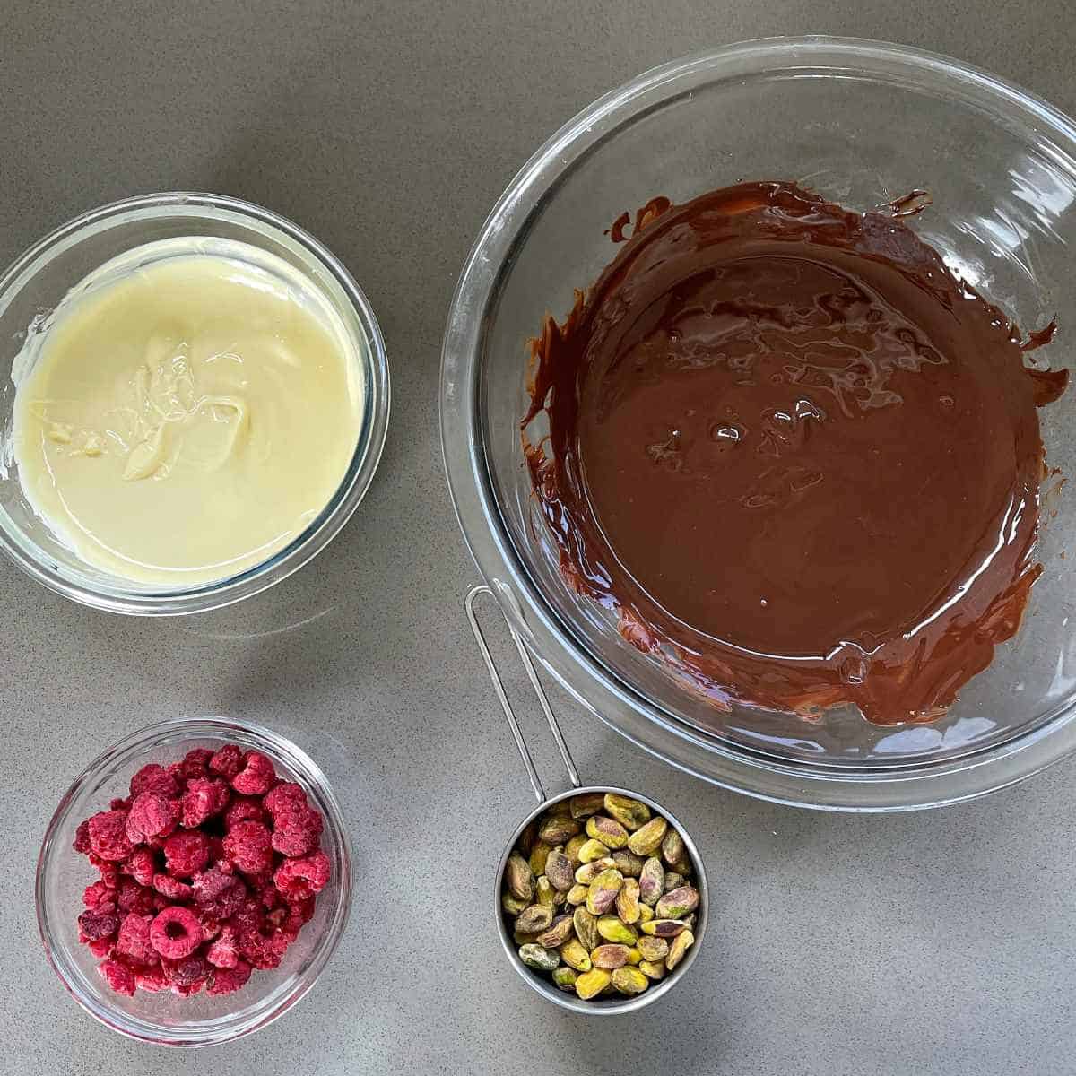 The melted milk and white chocolate and measured out raspberries and pistachios on a grey bench top for the chocolate bark.