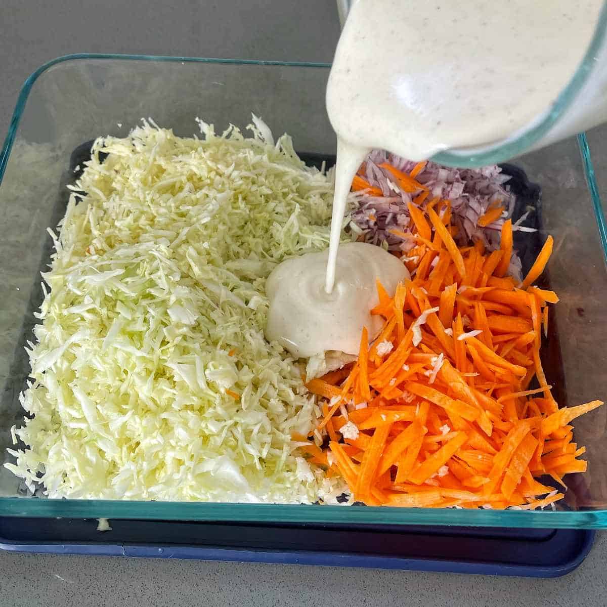 the sliced and grated ingredients for Coleslaw in a glass dish with the dressing being poured over the top.