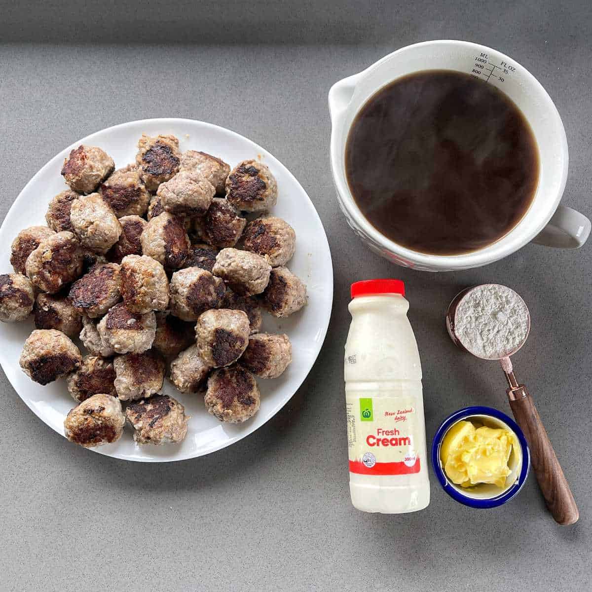 Cooked meatballs, gravy, cream, butter and flour all on a grey bench for assembling Swedish Meatballs.