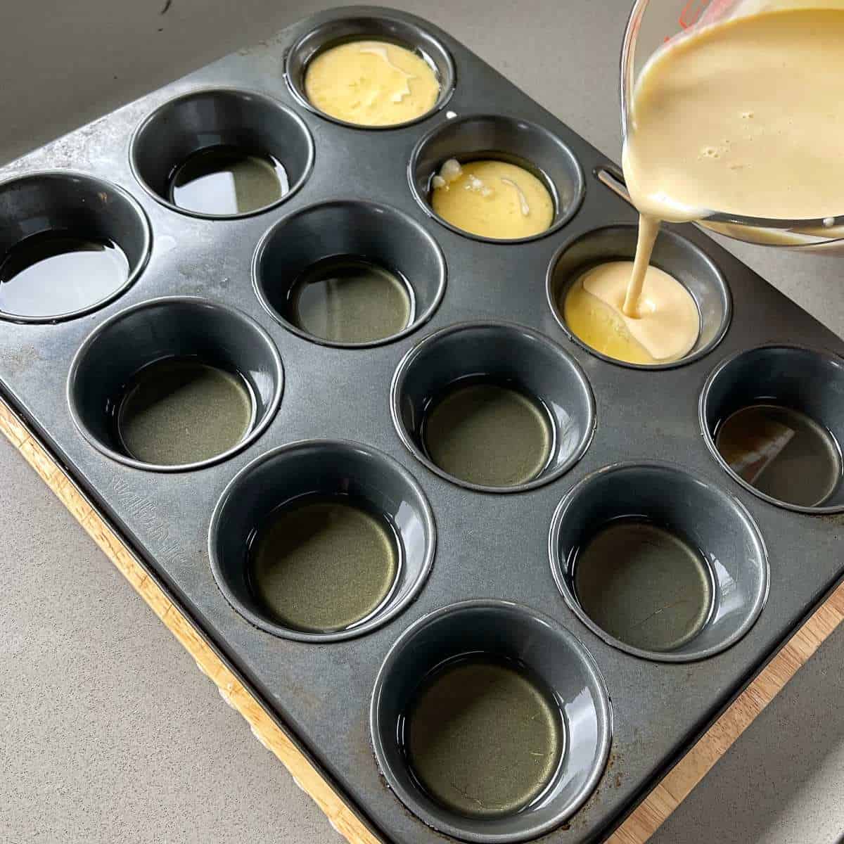 A glass jug with Yorkshire pudding batter pouring the mixture into individual portions in oiled muffin tin.