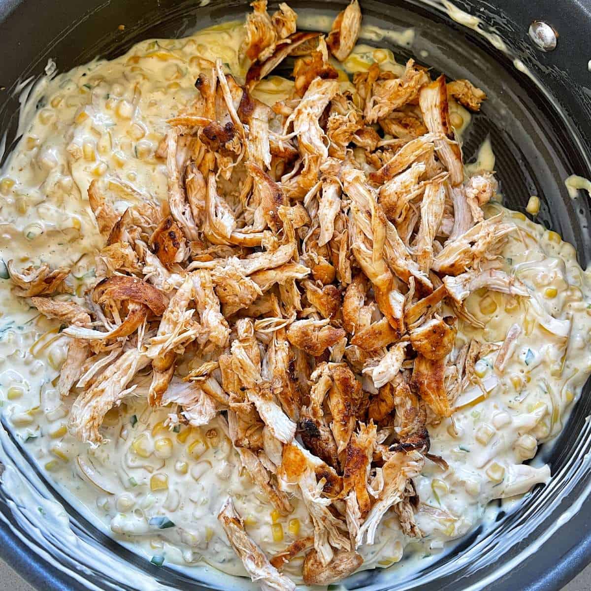 Shredded cooked chicken sitting on a creamy corn mixture in a frying pan. 