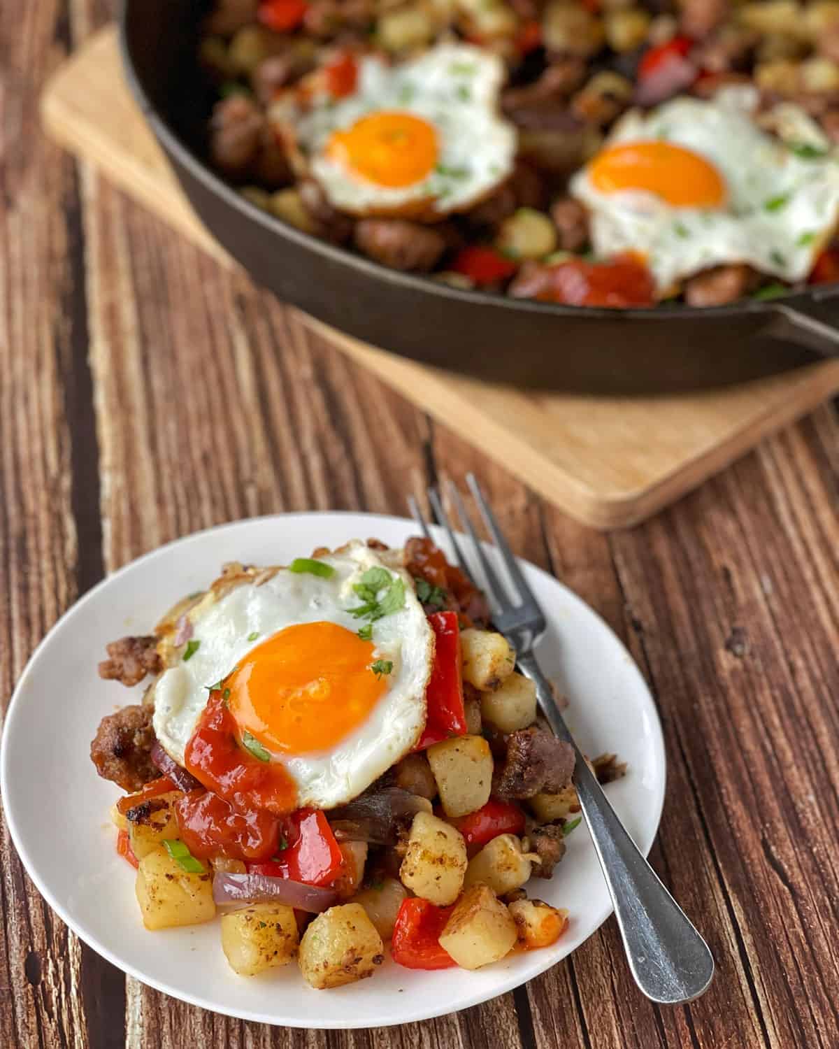 A serving of Skillet Sausage and Potatoes with a fried egg and tomato relish on a white plate with a fork resting on the side.