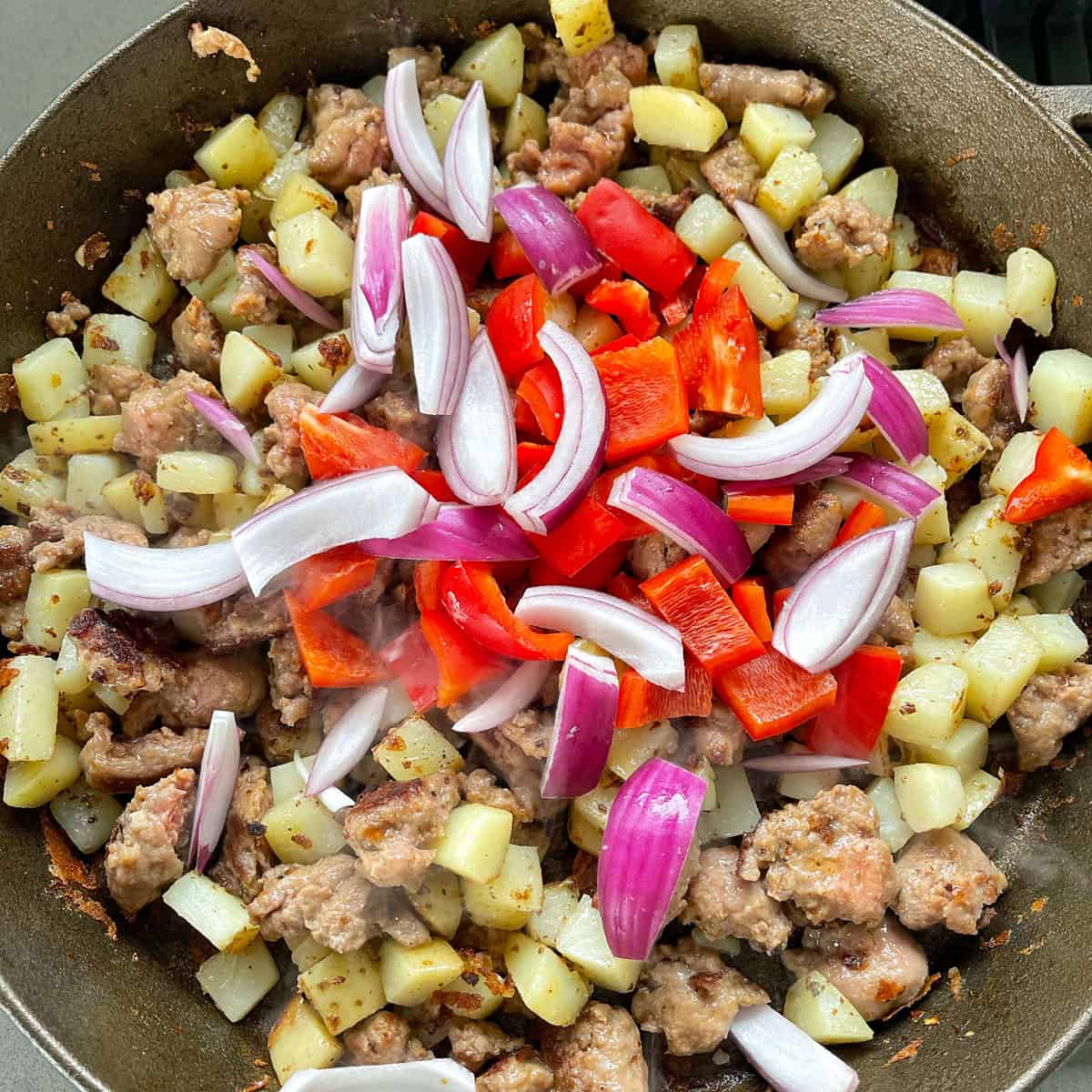 Fried potatoes and sausages in a large pan with fresh red onion and capsicum added.
