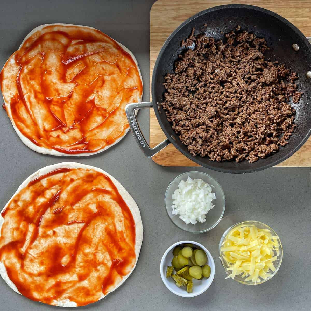 Two pizza bases with tomato sauce spread on them next to a frying pan with mince in it and three small bowls with cheese, onion and gherkins in them.