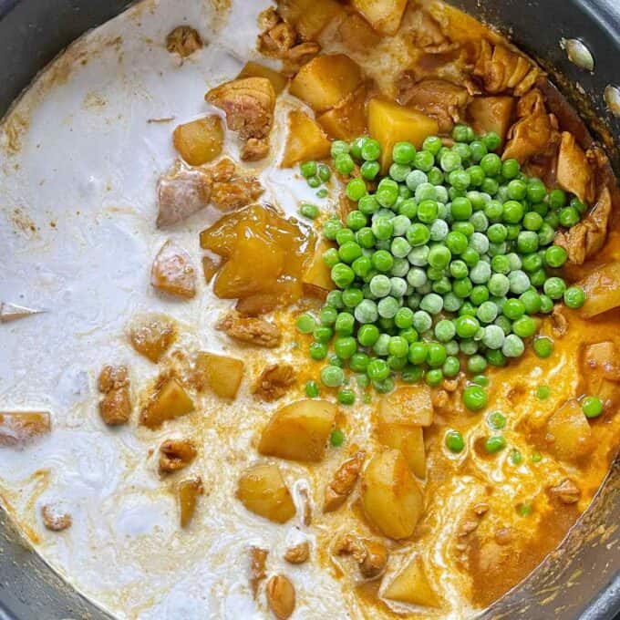 Cream and frozen peas being added to the chicken and potato curry mixture in a frypan.
