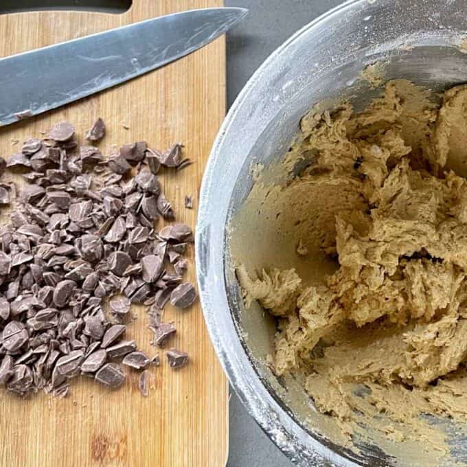 Sliced chocolate chips on a wooden chopping board with the cookie mixture in a bowl to the side.