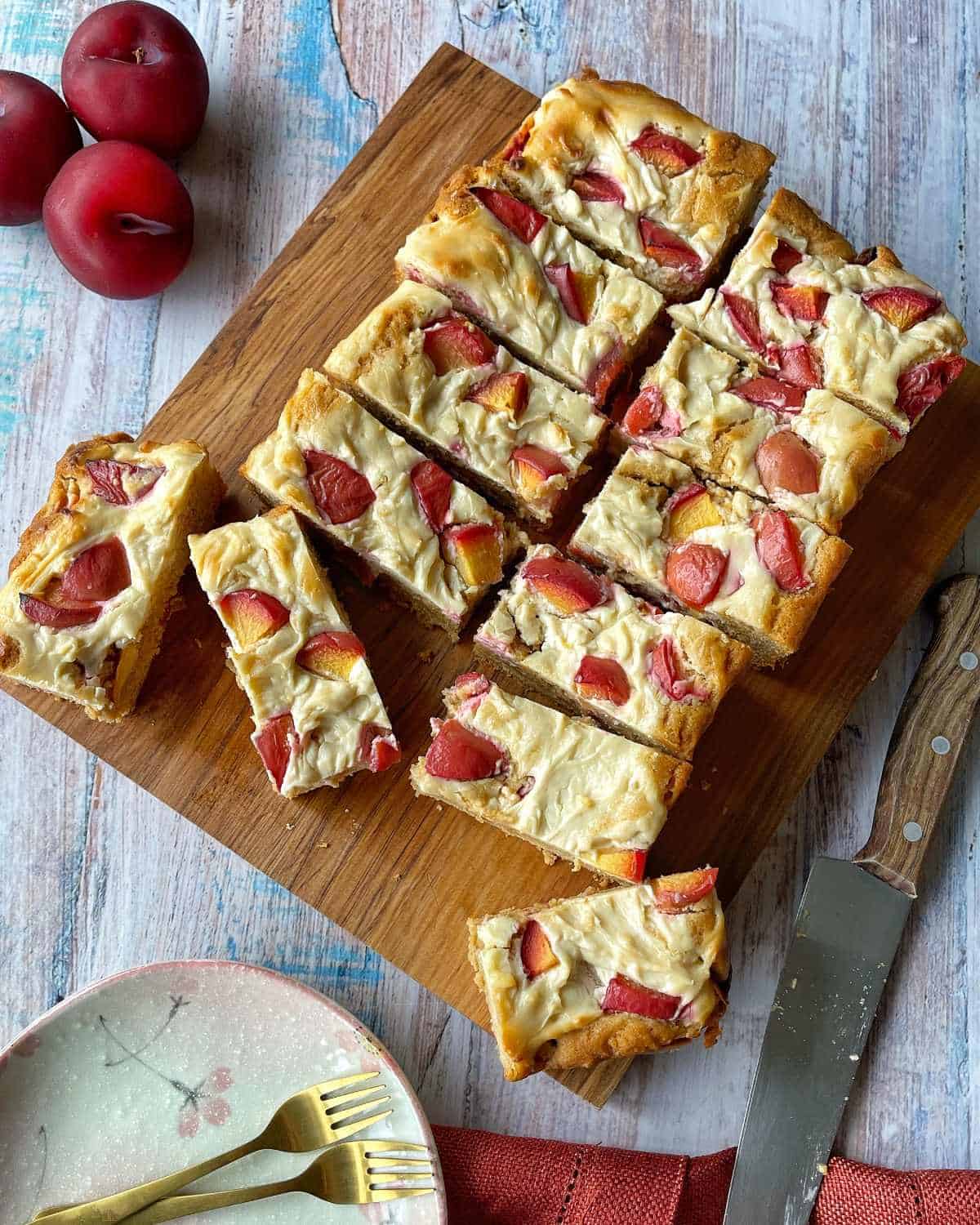 Cooked and sliced plum and cream cheese tray bake on a wooden chopping board.