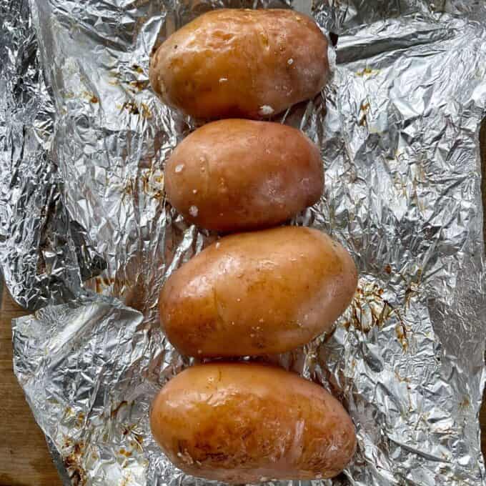 Four medium cooked red potatoes unwrapped from tinfoil on a wooden chopping board.