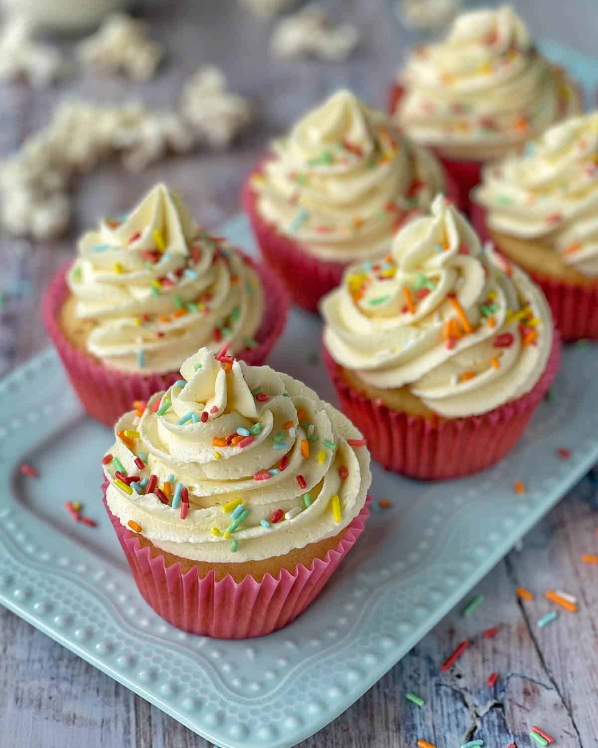 A close up shot of classic vanilla cupcakes with creamy butter icing and colourful sprinkles on top.