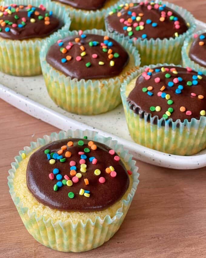 A close up shot of a classic vanilla with chocolate icing with colourful sprinkles on top. Several decorated cupcakes in the background on a ceramic plate.