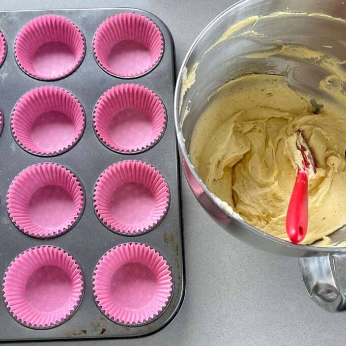 A birds eye view of a lined muffin tray with pink cupcake liners with the cupcake mixture in a mixing bowl sitting on a grey bench.
