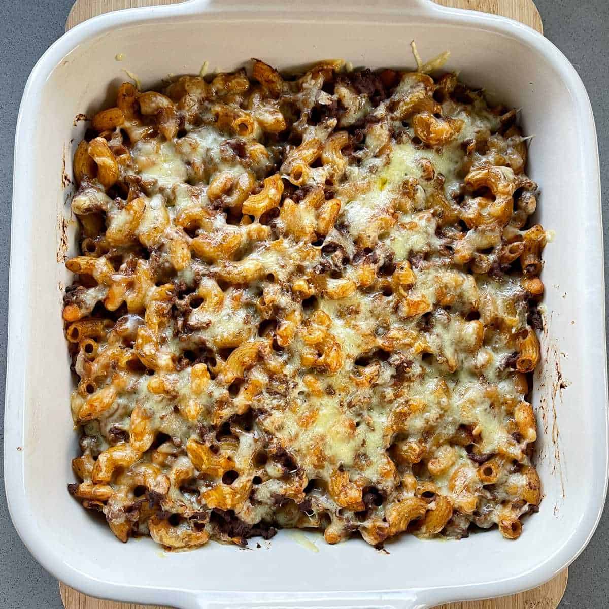 A baked mince, macaroni and bean casserole on a wooden chopping board.