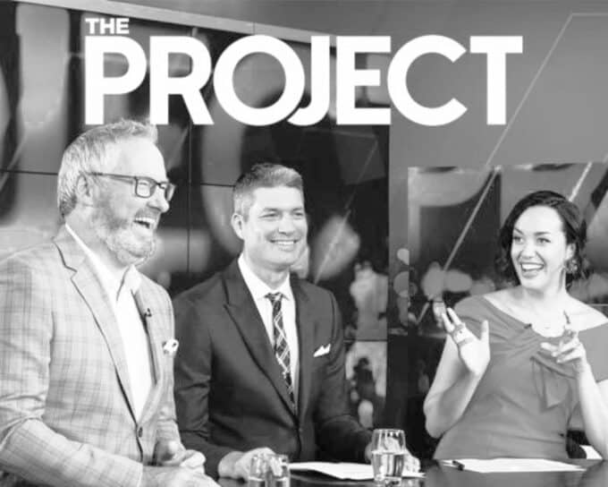 A black and white photo of two men and one woman sitting at a news desk with the words 'The Project' above them.