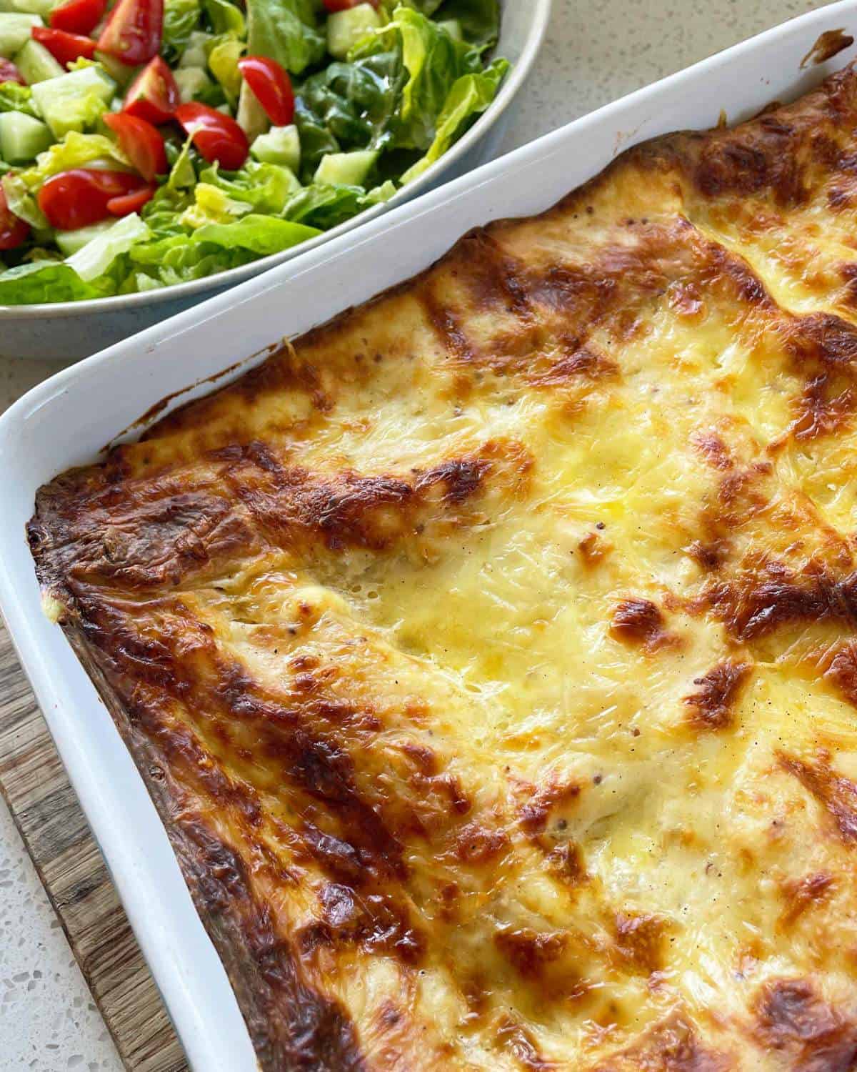 A cooked Chicken Lasagne sitting on a wooden bench, a green salad to the side.