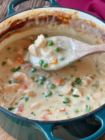Creamy chicken casserole served in the ceramic dish it was made in sitting on a wooden chopping board with a wooden spoon showing the creamy chicken and vegetable filling.