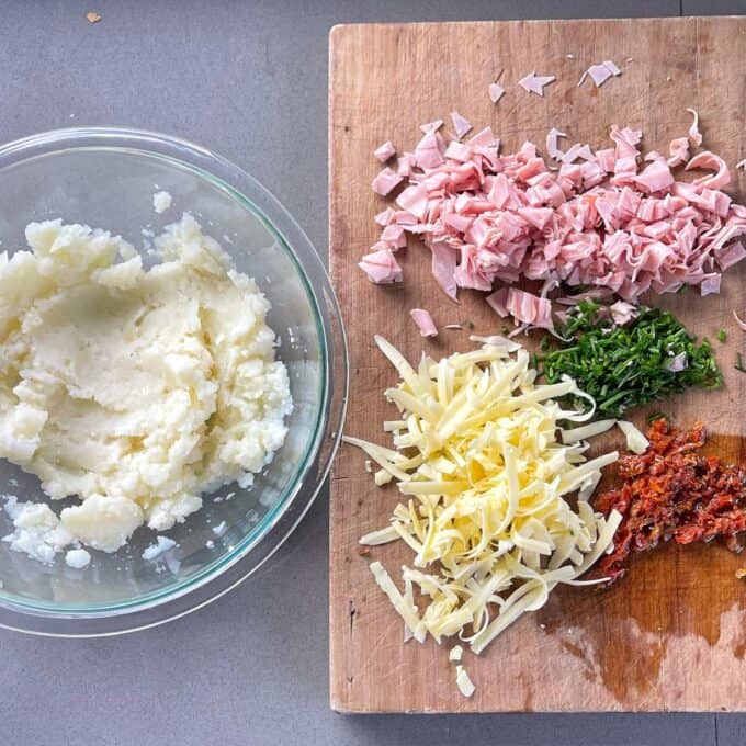 Sliced ham, chives, sundried tomatoes and grated cheese on a wooden chopping board. Mashed potatoes in a glass bowl,