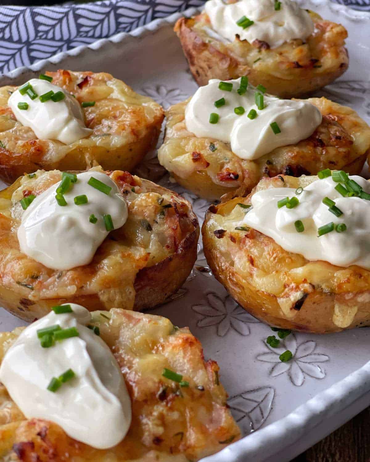 Six cooked Jacket Potatoes filled with ham and cheese, served with sour cream and chives on a white platter.