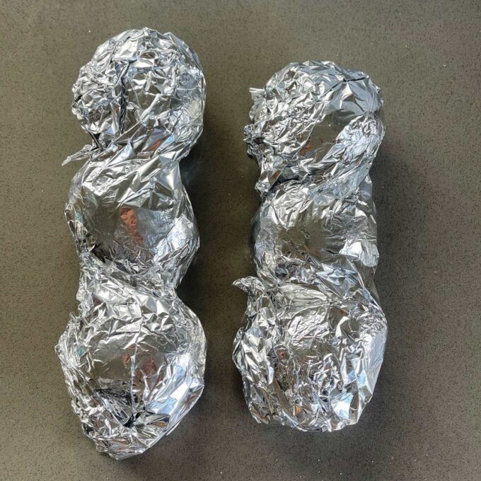 Six potatoes wrapped in tinfoil on a grey bench top.
