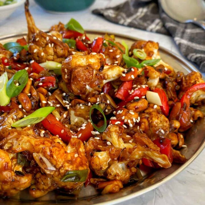 A close up of Kung Pao Cauliflower served in a large bowl with sliced spring onion and sesame seeds scattered over the top.