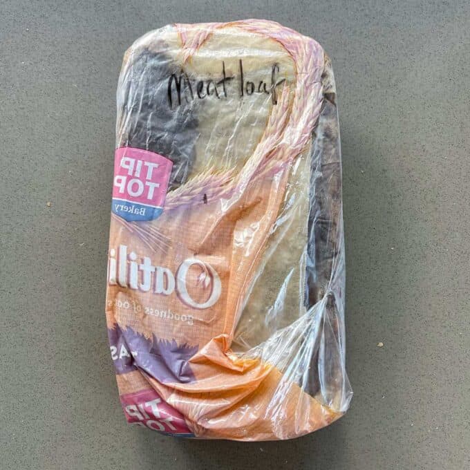 An uncooked meatloaf, wrapped in baking paper, inside a bread bag with meatloaf written on it, sitting on a grey bench top. 