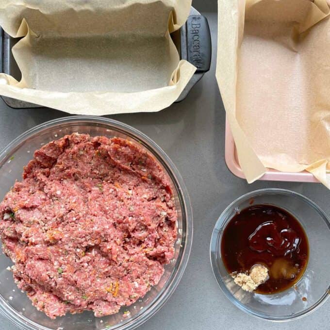 Two lined loaf tins with baking paper, the combined meat mixture and the sauce all sitting on a grey bench top about to be assembled.
