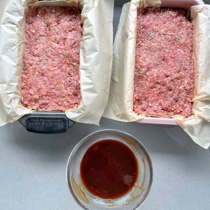 Two lined loaf tins with baking paper, with combined meat mixture and the sauce all sitting on a grey bench top. 