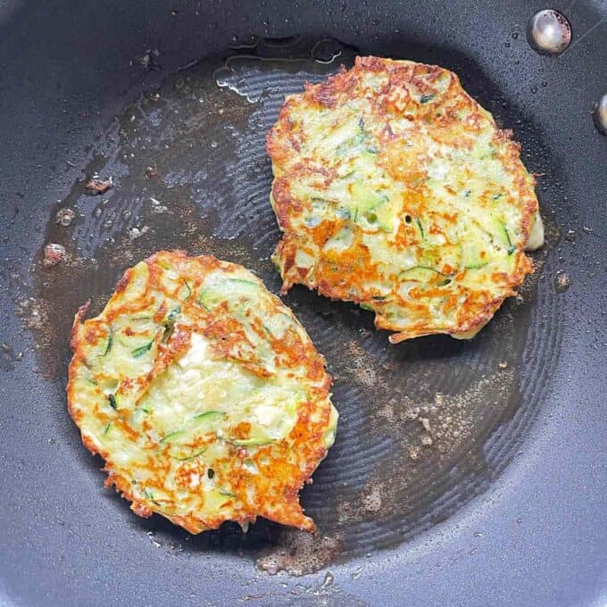 Two courgette and feta fritters frying a non-stick frying pan.