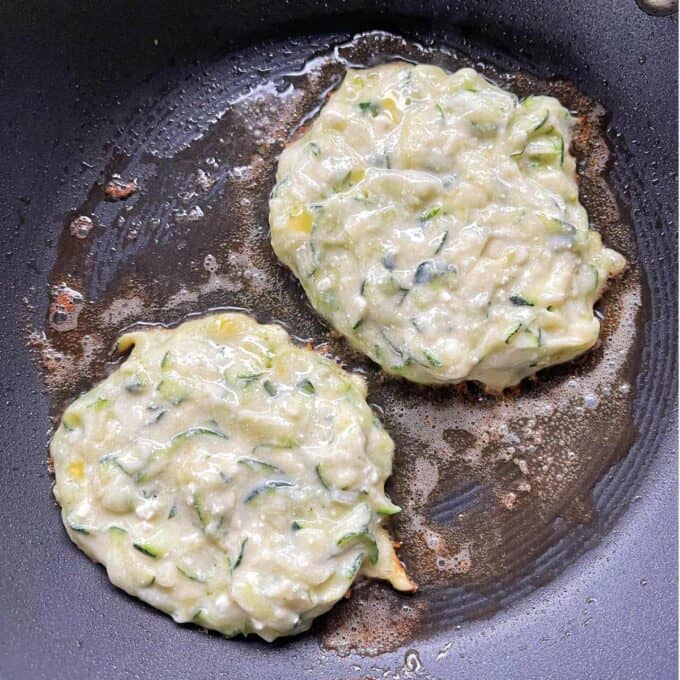 Two courgette and feta fritters frying a non-stick frying pan.