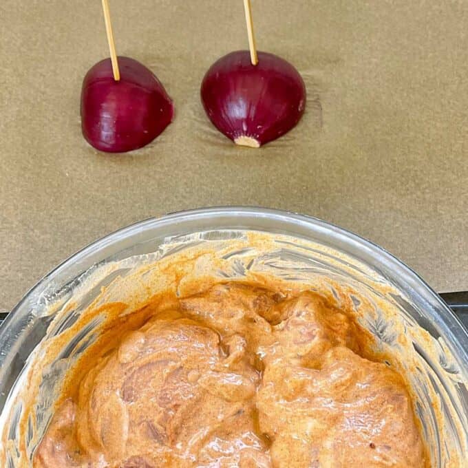 A glass bowl with marinaded chicken and a red onion cut in half, with a wooden skewer in each piece of onion. 