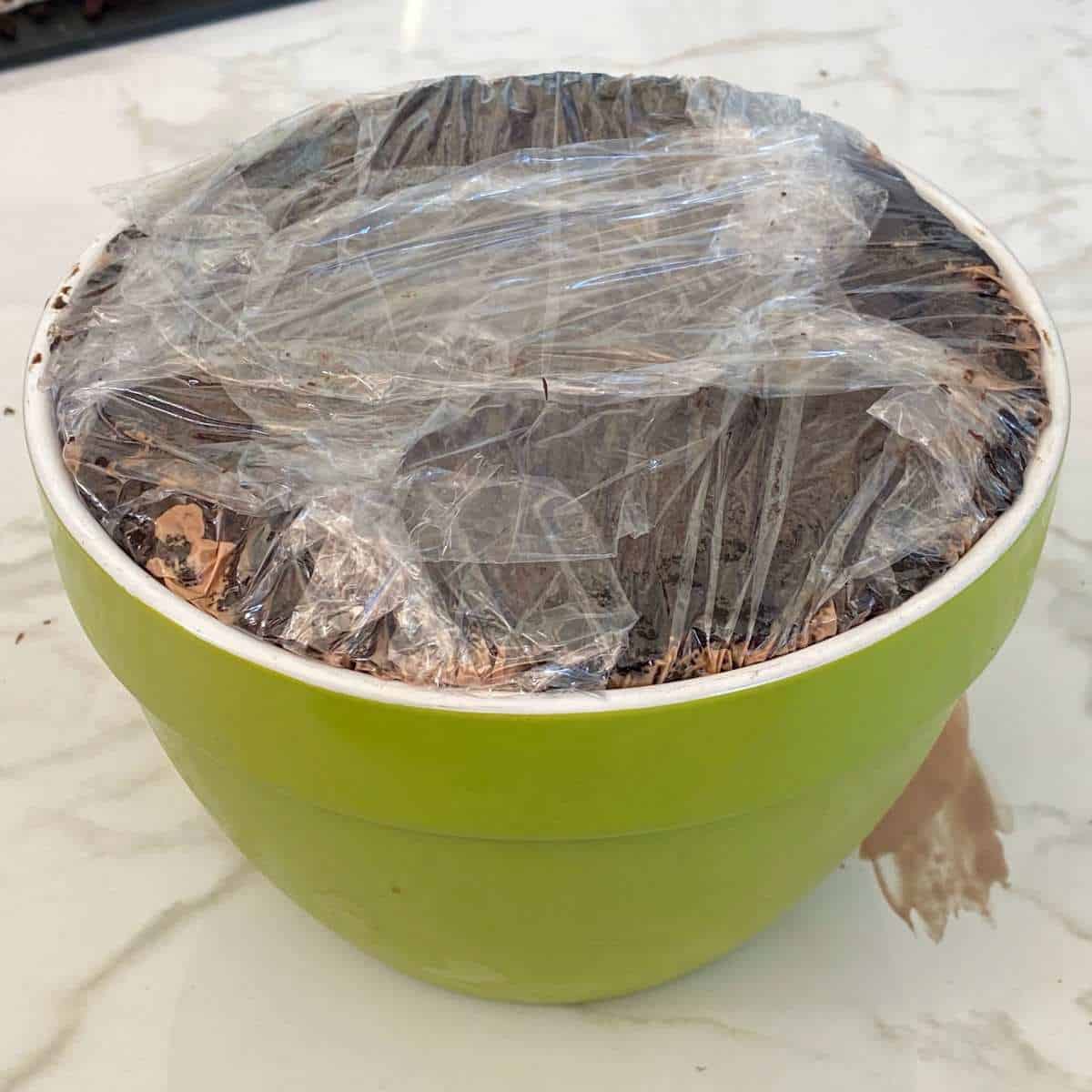 A green bowl filled with ice cream and cake covered with cling film on a marble bench.