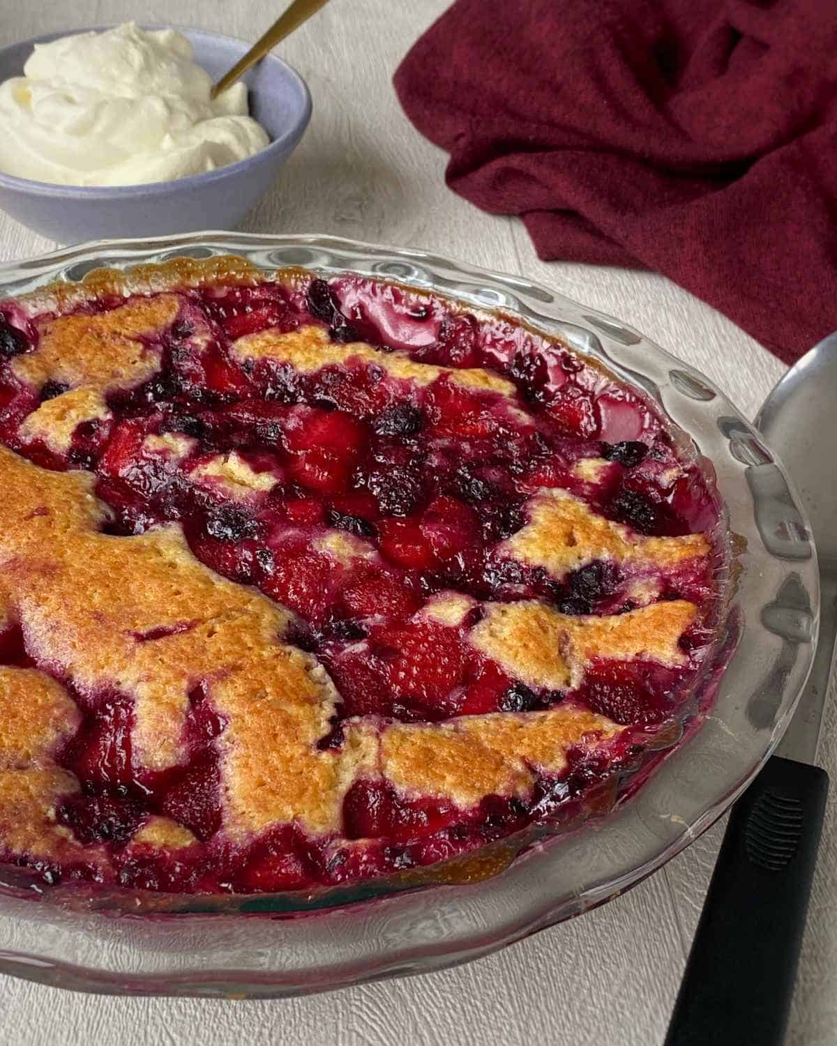 A close up shot of Berry Self Saucing Pudding in a round glass pie dish with a small bowl of whipped cream in the background.