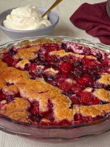 A close up shot of Berry Self Saucing Pudding in a round glass pie dish with a small bowl of whipped cream in the background.