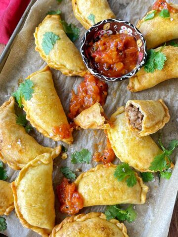 A selection of cooked cheesy beef empanadas with pasley scattered over the top and a chutney in a small dish for dipping.