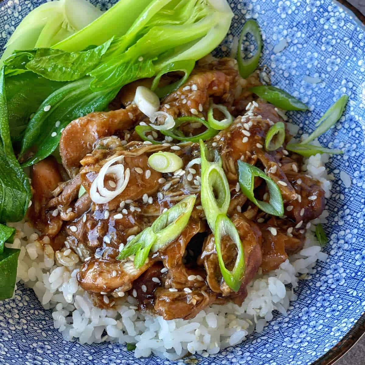 Honey Soy Chicken served over rice in a blue bowl with bok choiy and sliced spring onion and sesame seeds scattered over the top.